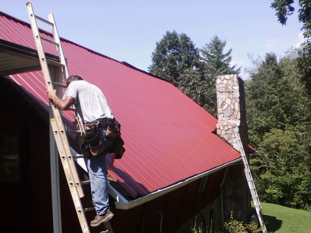 Roofer on a ladder beside a red metal roof and stone chimney — Soffit & Fascia Repair by Roofing Brothers in Montezuma